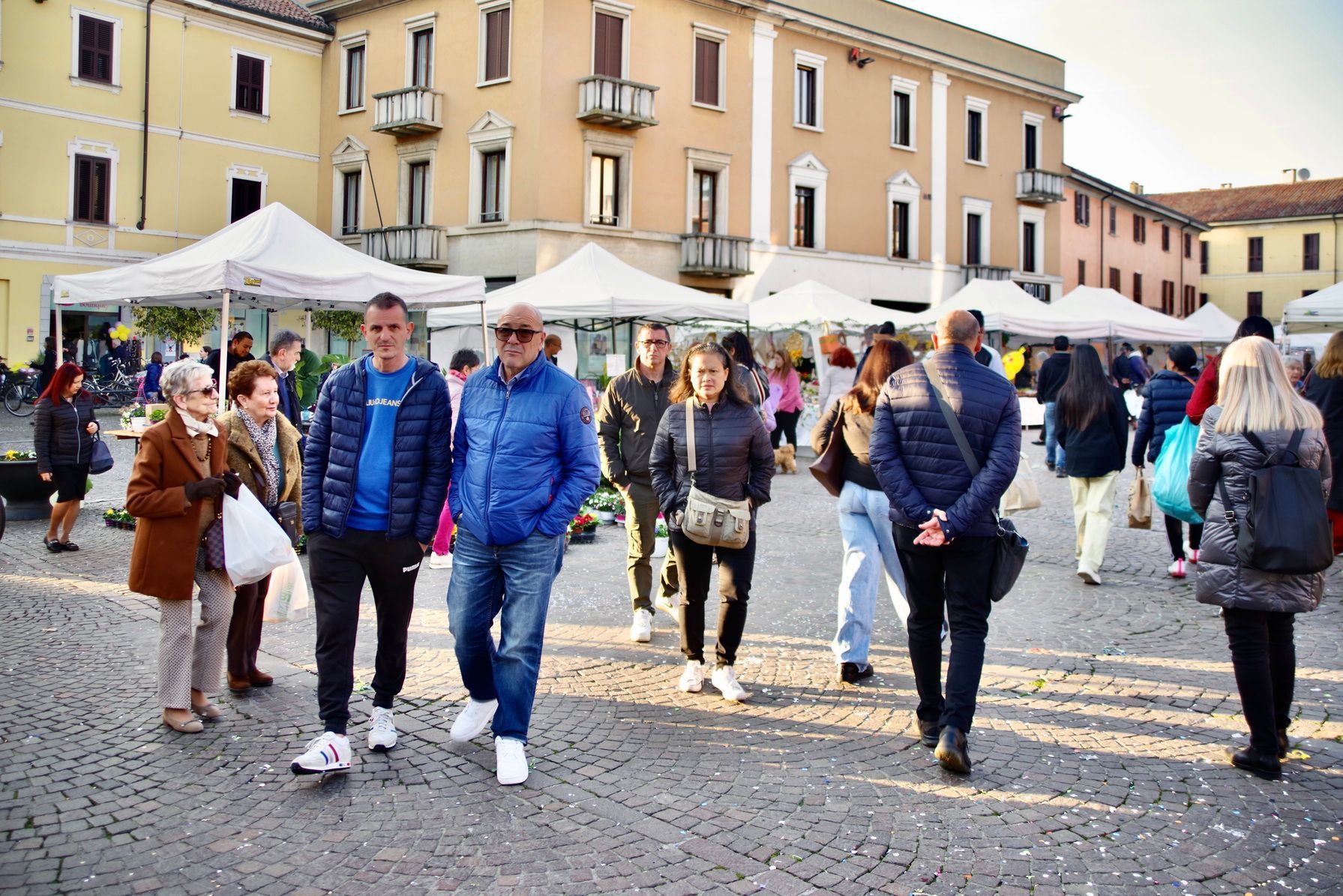 Persone che camminano in una piazza cittadina con tende bianche e edifici sullo sfondo.