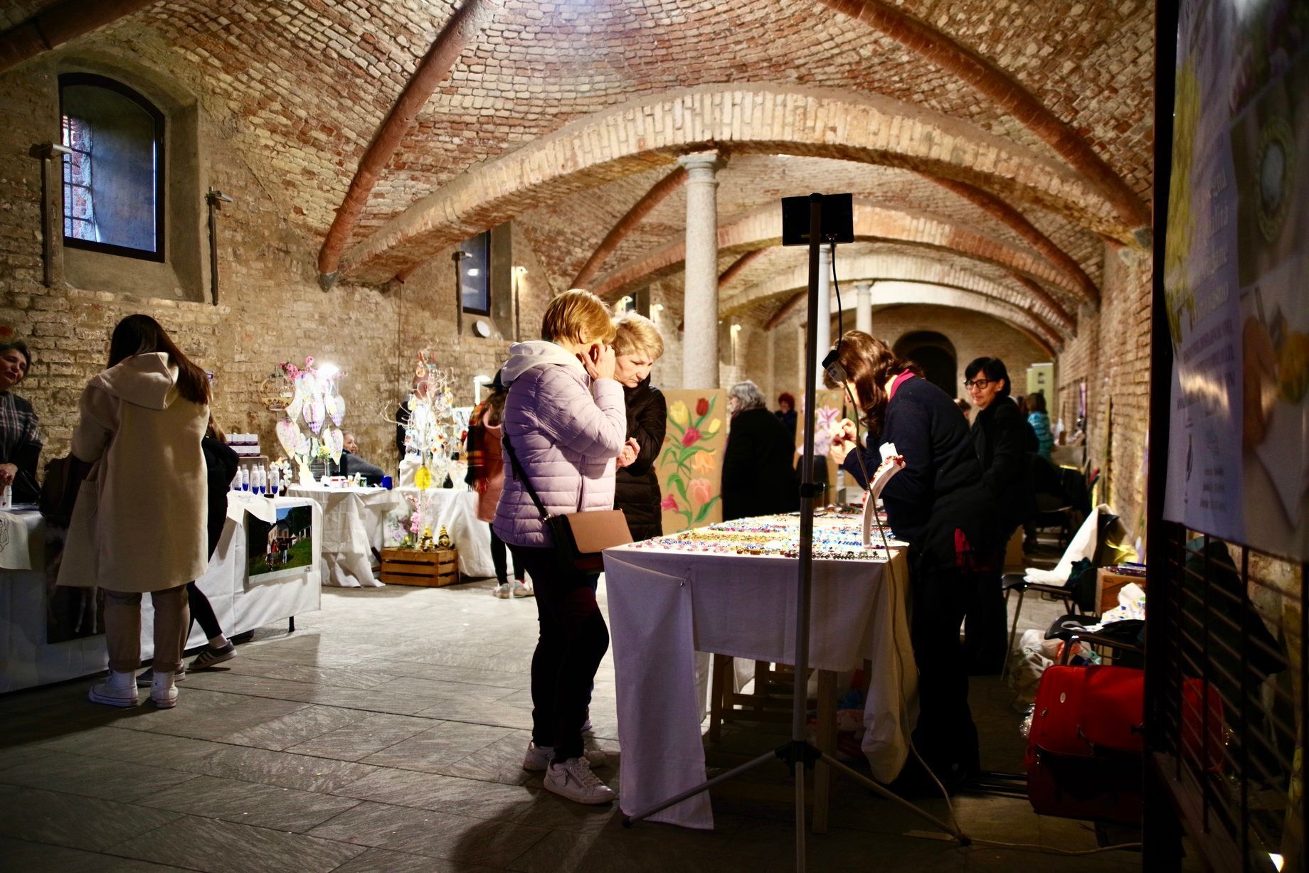 La gente curiosa tra i banchi dei venditori in un mercato coperto. Soffitto in mattoni, luci soffuse.
