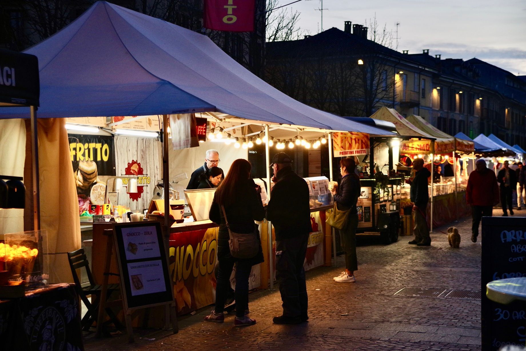 Bancarelle di cibo in un mercato notturno, gente che curiosa, venditori che servono, sotto tendoni bianchi e lucine.