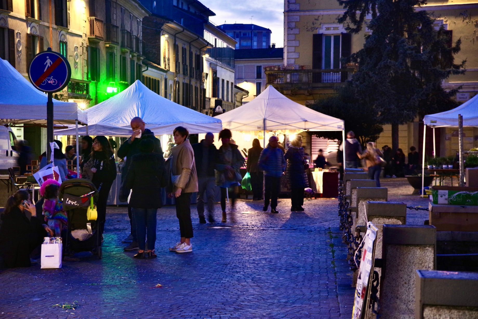 Persone al mercato sotto tende bianche in una piazza europea al tramonto.