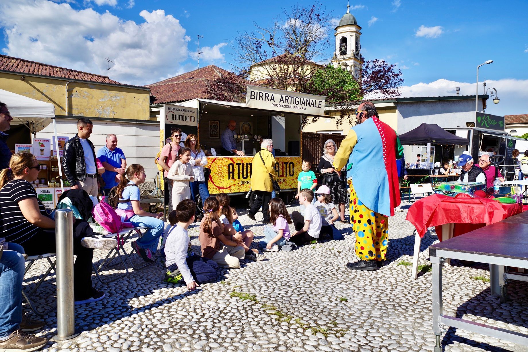 Un clown intrattiene i bambini in un mercato all'aperto con venditori di cibo, in una giornata di sole.
