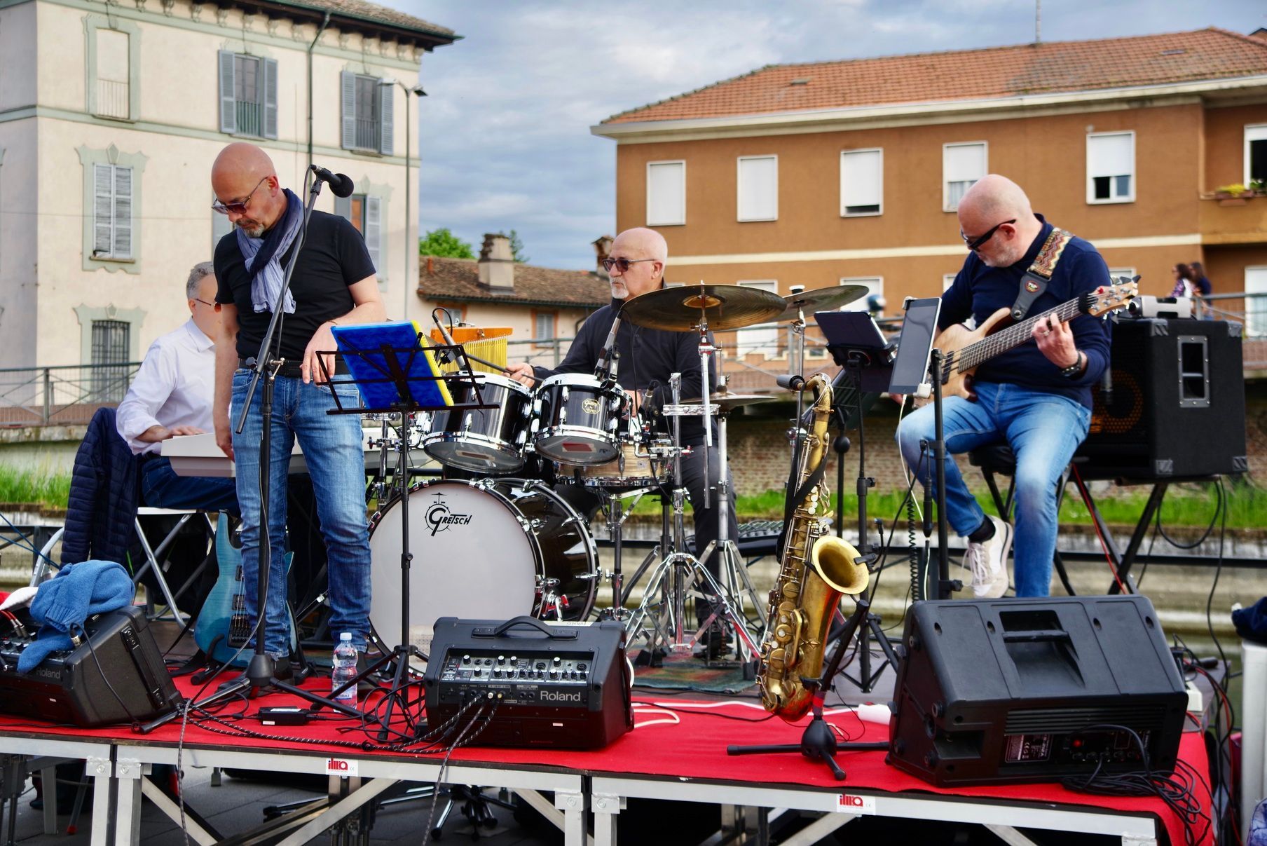 Un gruppo jazz si esibisce all'aperto su un palco. Quattro uomini calvi suonano batteria, chitarra, sassofono e tastiere. Edifici in mattoni sullo sfondo.