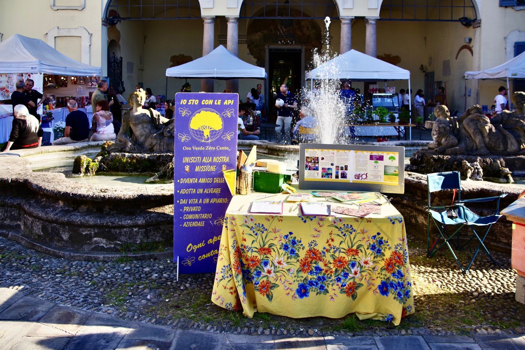 Fiera all'aperto con stand, una fontana e persone, con un tavolo coperto da una tovaglia floreale.