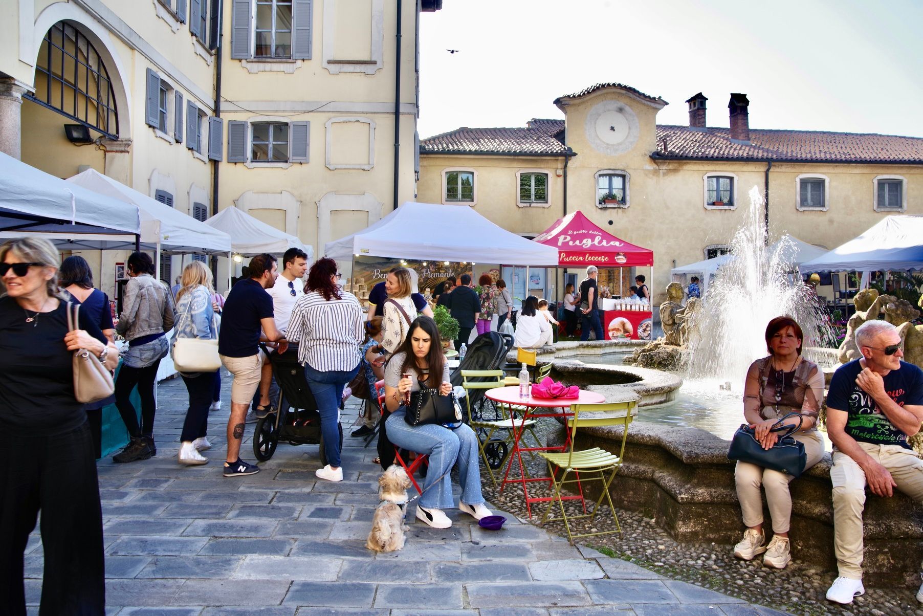 Persone in un mercato all'aperto con tende, una fontana ed edifici in una piazza italiana.