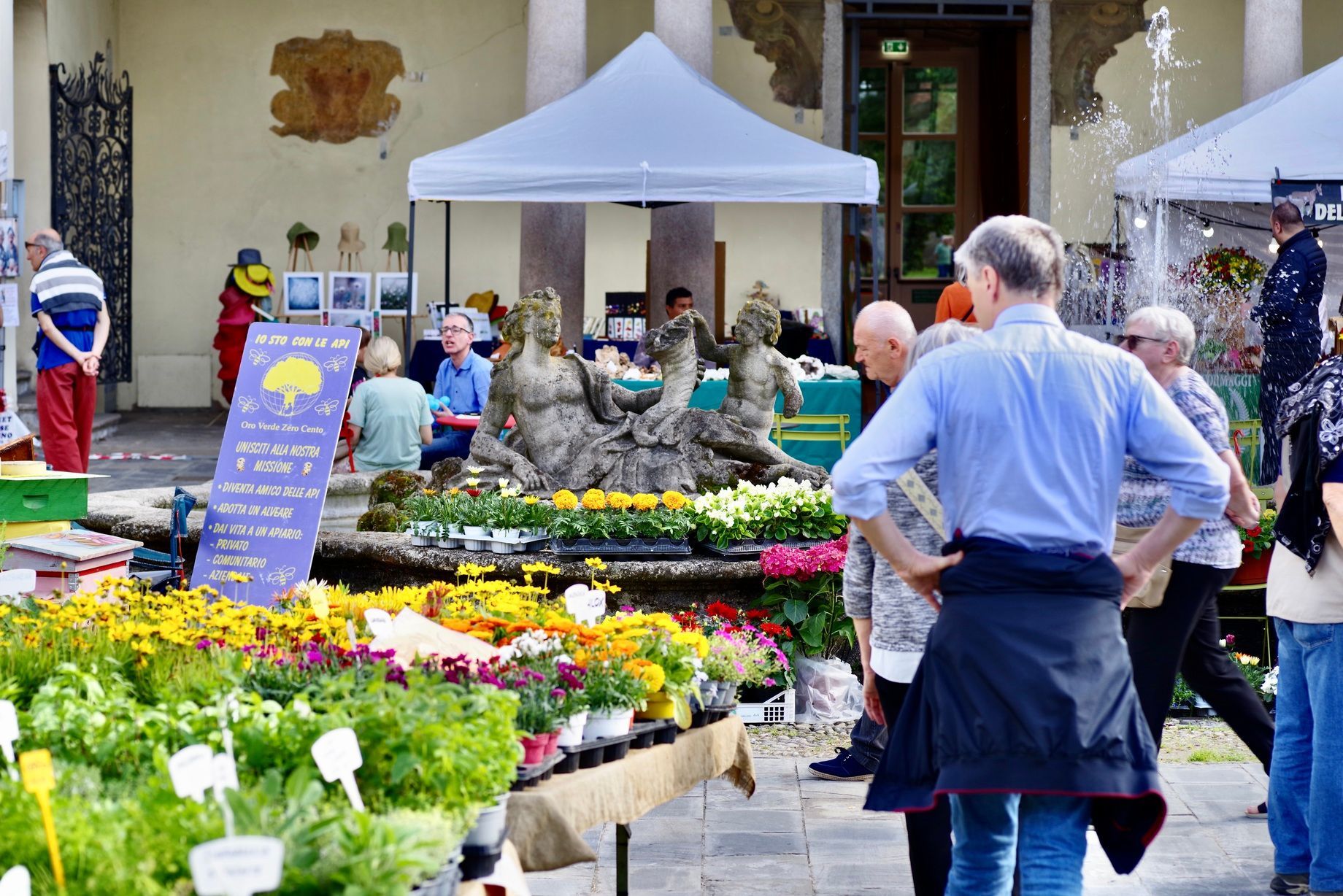 La gente curiosa tra le esposizioni di fiori e piante in un mercato all'aperto vicino a una fontana con sculture; giornata di sole.