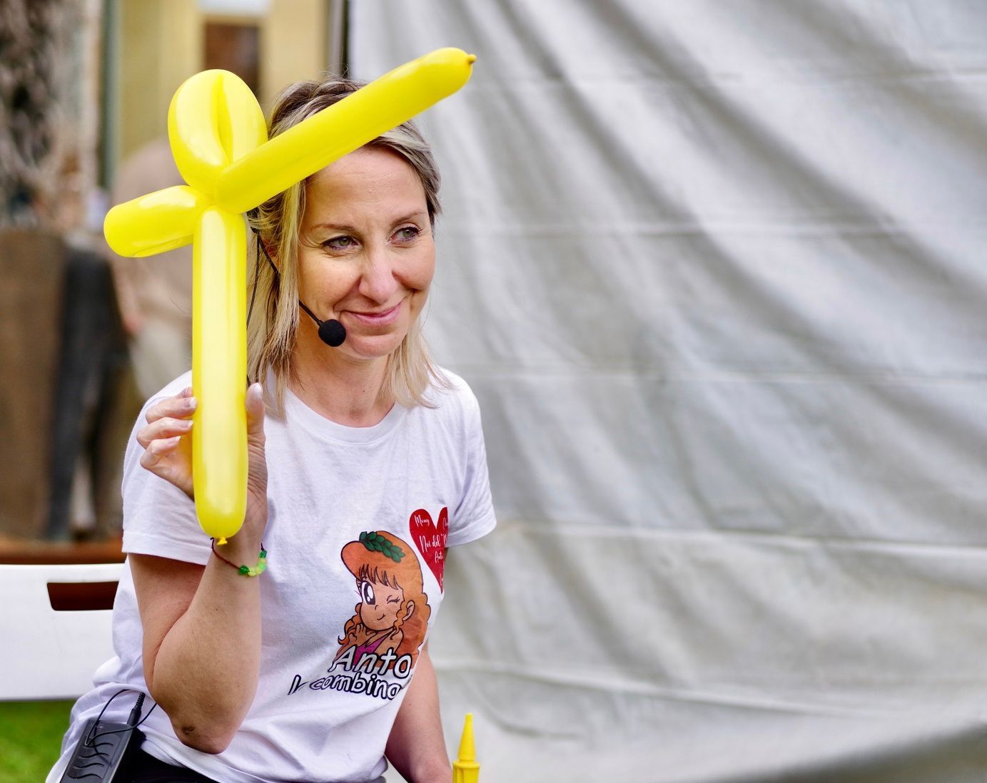 Donna che tiene in mano un palloncino giallo a forma di croce, sorridente, con indosso una maglietta bianca, all'aperto.