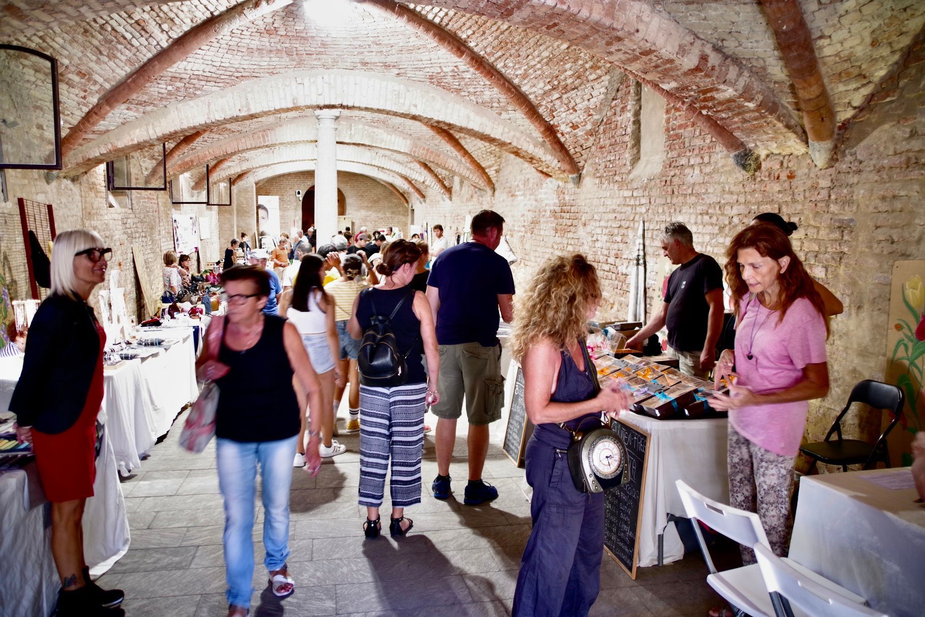 Persone che curiosano tra i prodotti artigianali in un mercato coperto, sotto soffitti a volta in mattoni.