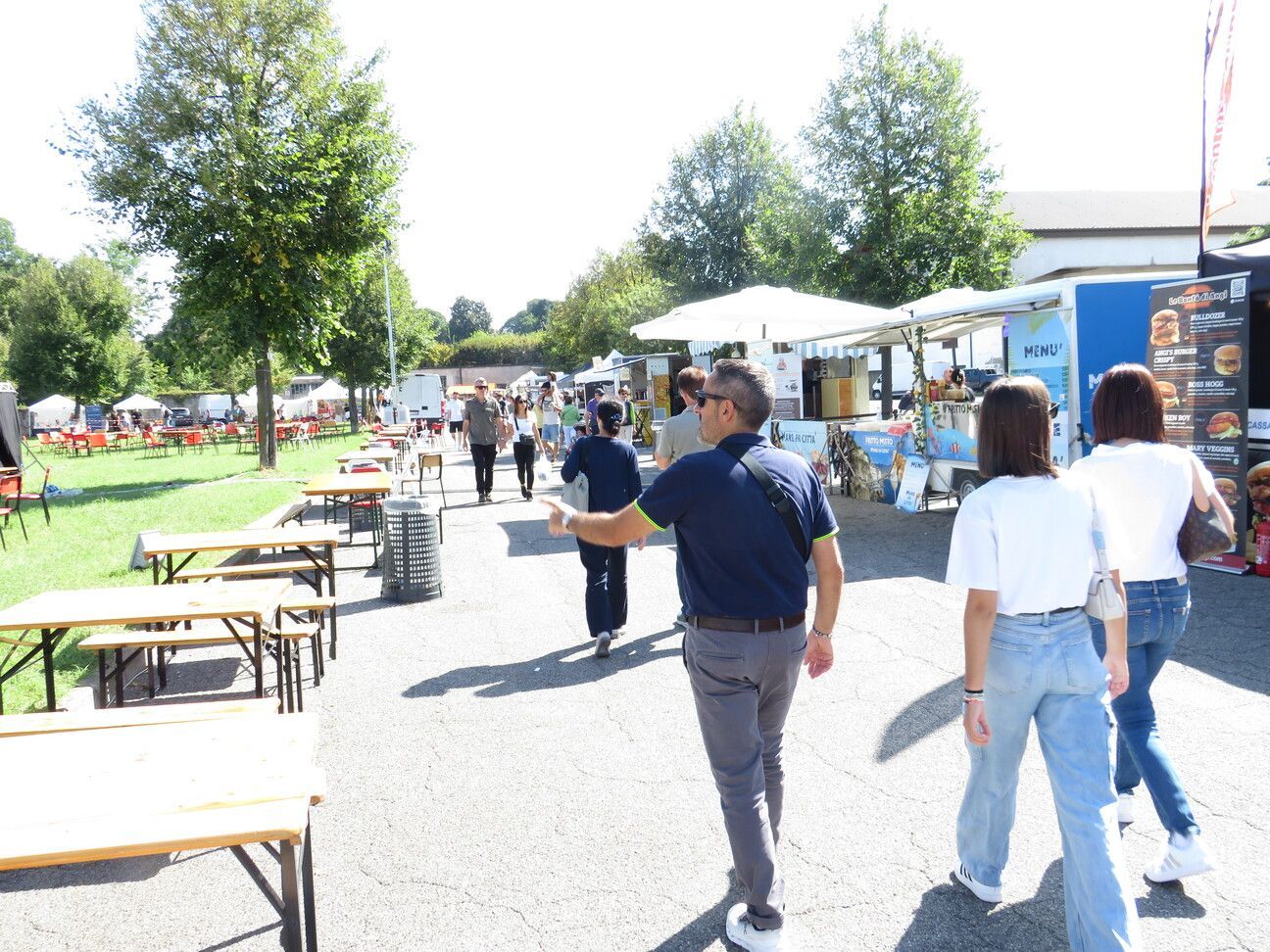 Persone che camminano durante un festival gastronomico. Food truck e tavoli da picnic sono visibili sotto un cielo soleggiato.