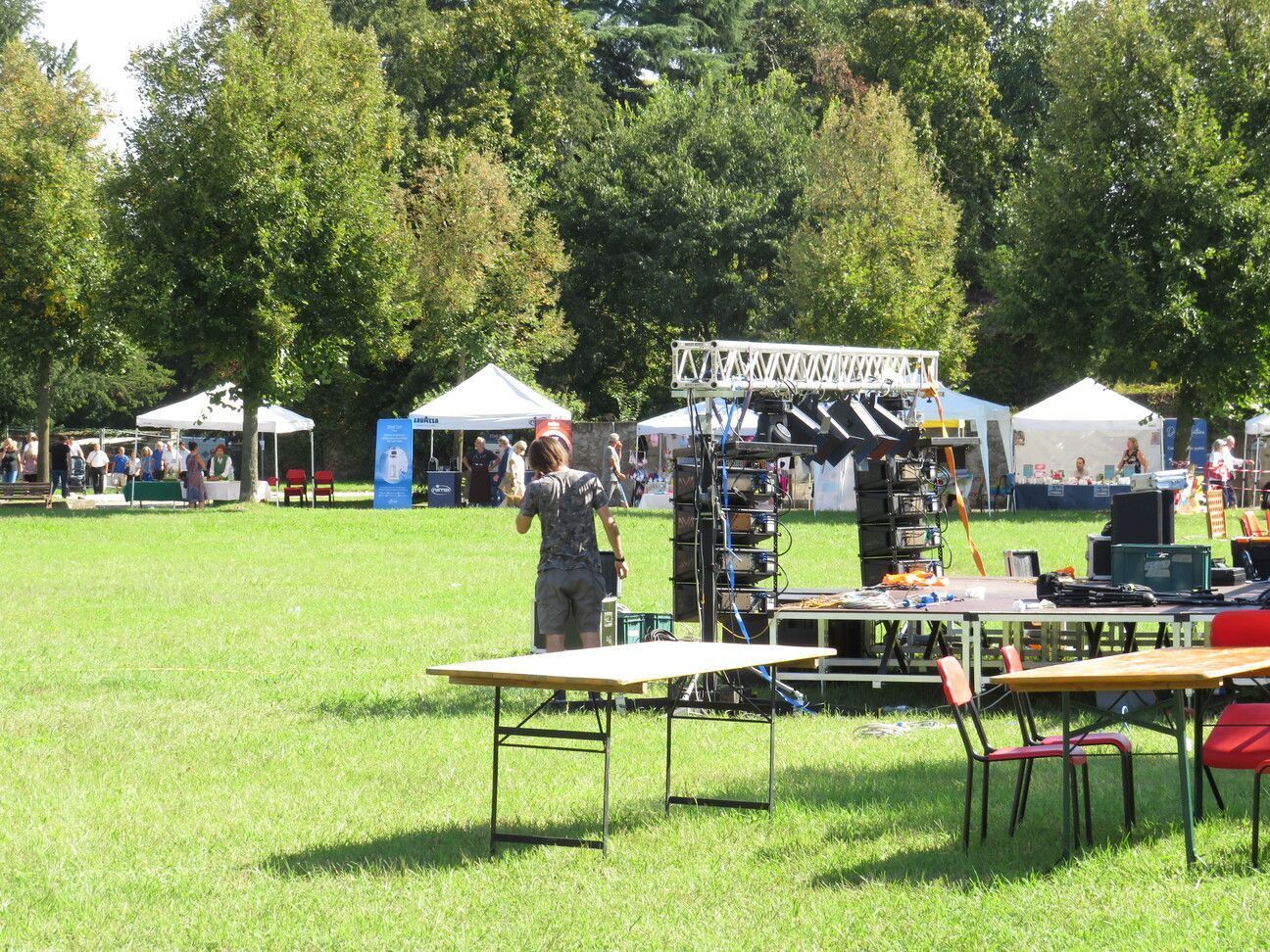 Scena di un parco con tavoli, tende, una persona che installa l'attrezzatura audio e alberi.