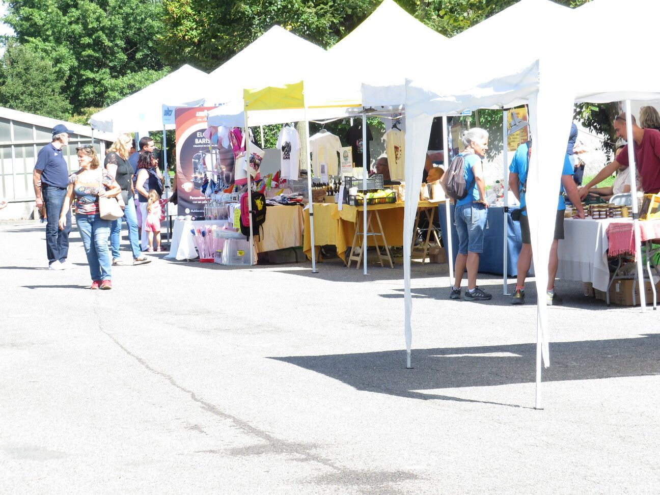 Mercato all'aperto con tende bianche, persone che curiosano tra i banchi con la merce in una giornata di sole.