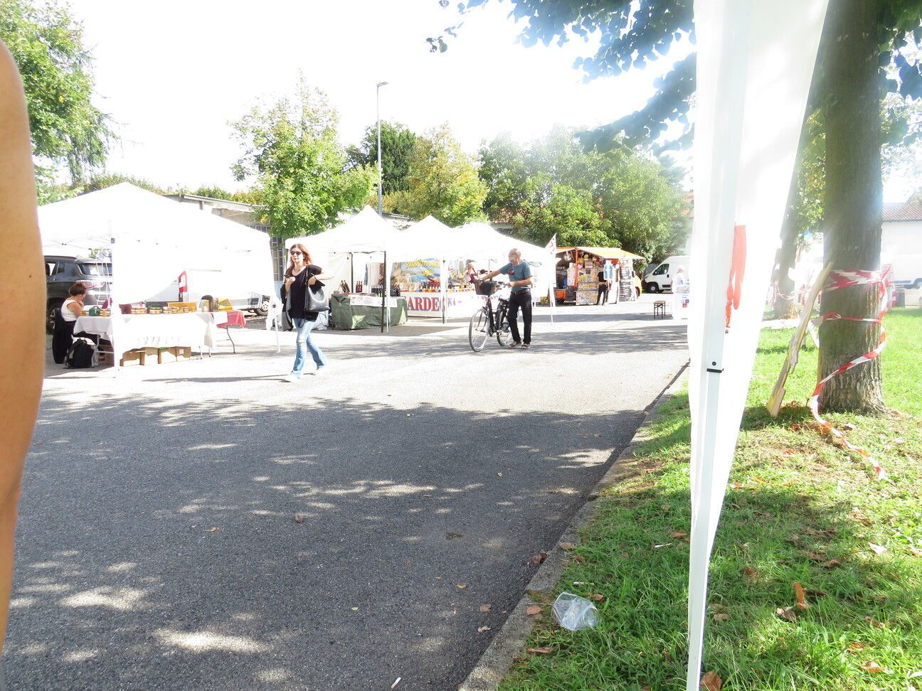 Mercato all'aperto con tende bianche, gente che cammina ed erba. Giornata di sole.