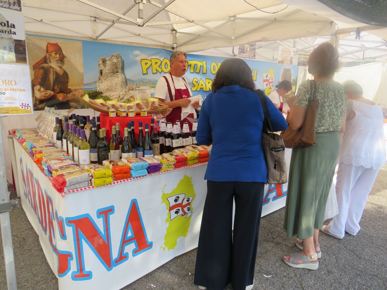 Un venditore al mercato sardo vende vino, marmellate e formaggi a tre clienti.