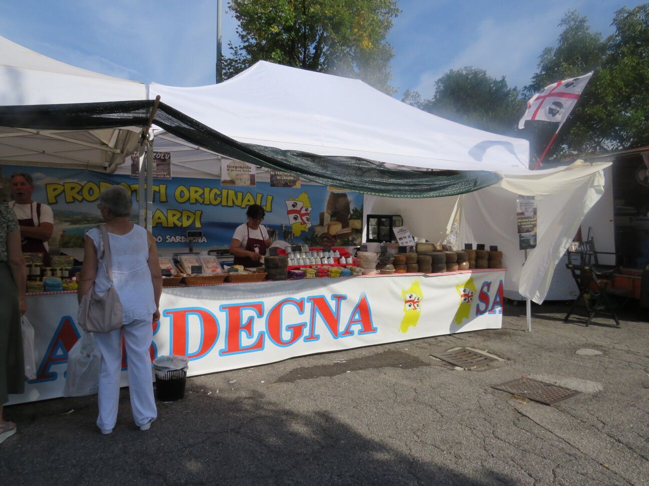 Uno stand gastronomico sardo con prodotti esposti, persone che guardano e una bandiera.