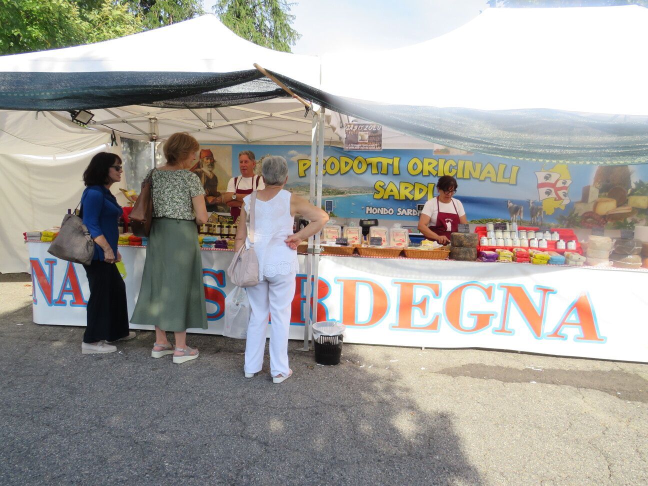 Bancarella del mercato: persone che curiosano tra i prodotti sardi. Tenda bianca, prodotti colorati, striscione con la scritta 