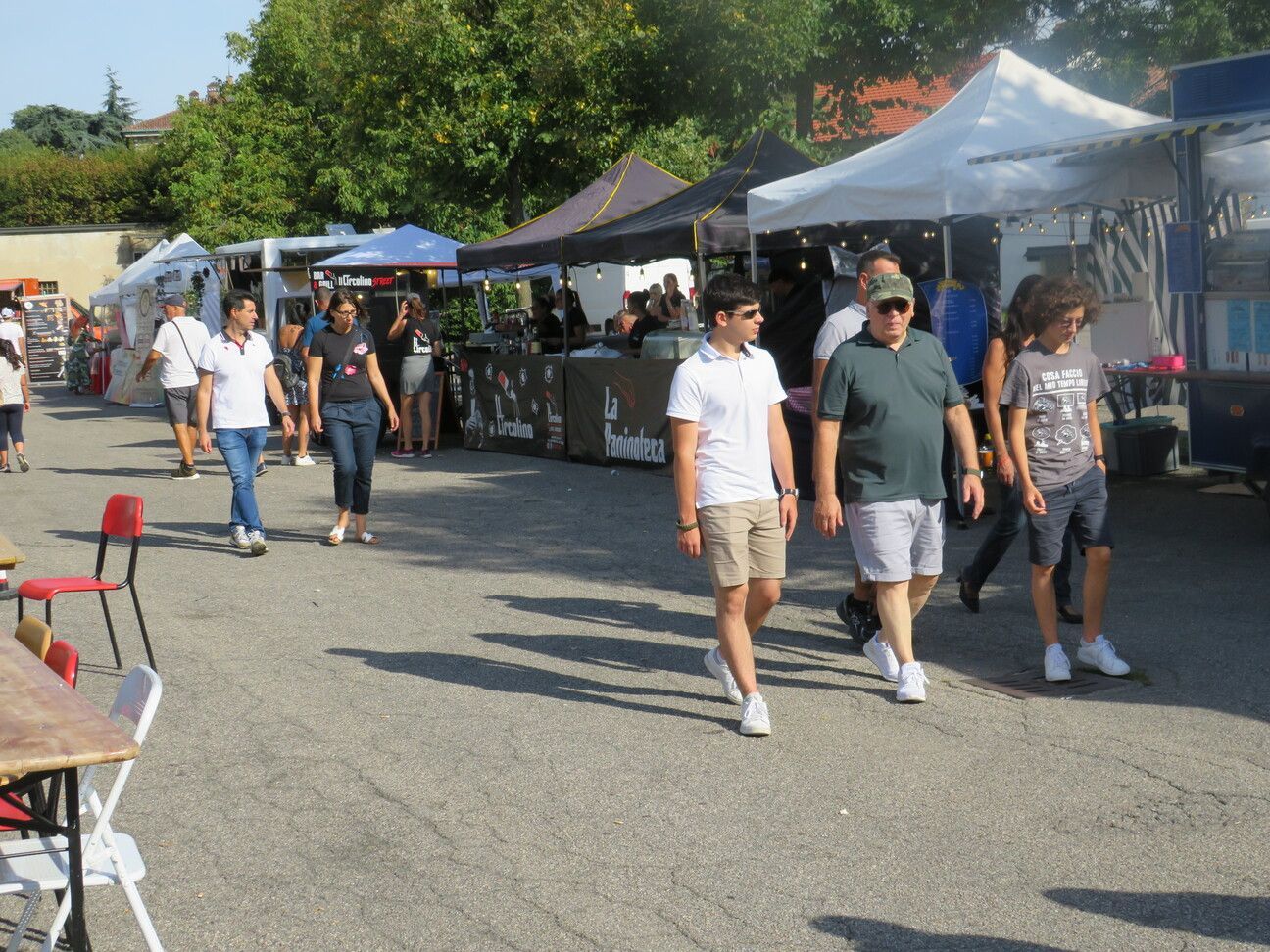 Persone che camminano tra le tende dei venditori in un mercato all'aperto in una giornata di sole.