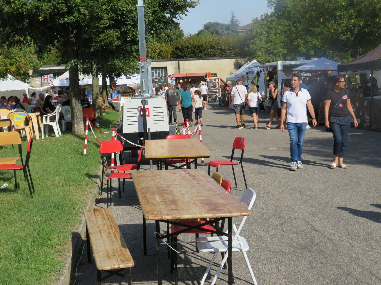 Scena di un mercato alimentare all'aperto: tavoli, sedie, persone che camminano, bancarelle dei venditori, alberi e luce solare intensa.