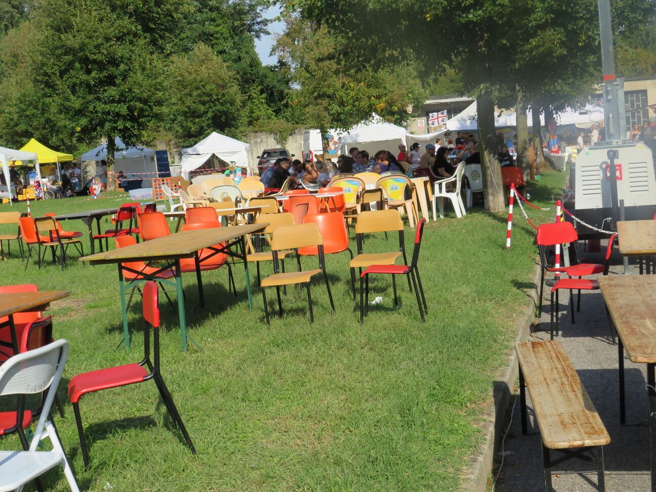 Evento all'aperto con tavoli e sedie sull'erba; persone sullo sfondo.
