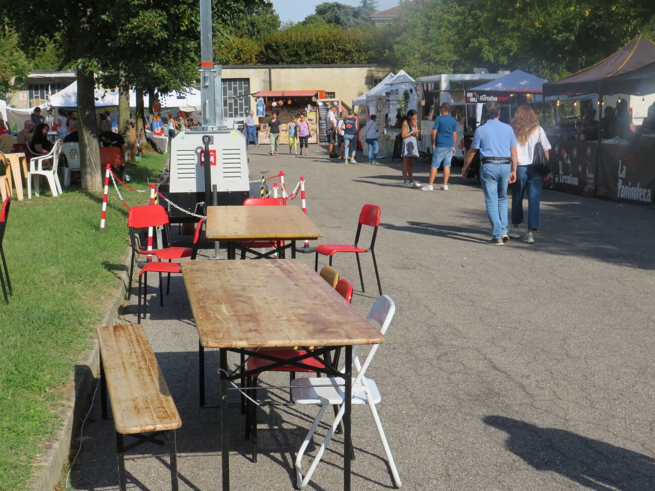Scena di mercato all'aperto: tavoli e sedie in primo piano, persone che curiosano tra le bancarelle sullo sfondo.