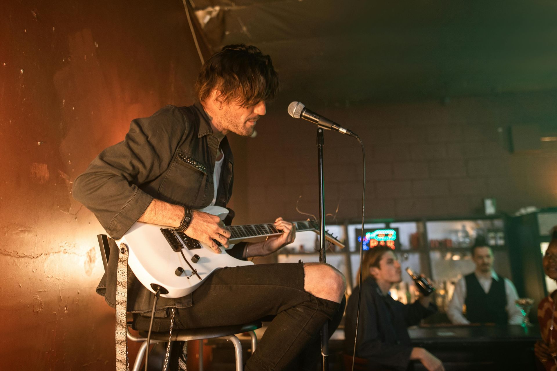 Un uomo suona una chitarra elettrica bianca e canta in un microfono in un bar scarsamente illuminato.