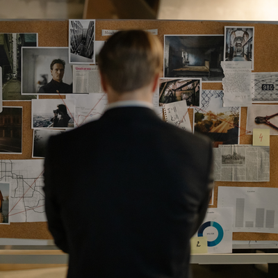 A man in a suit stands in front of a bulletin board with pictures on it