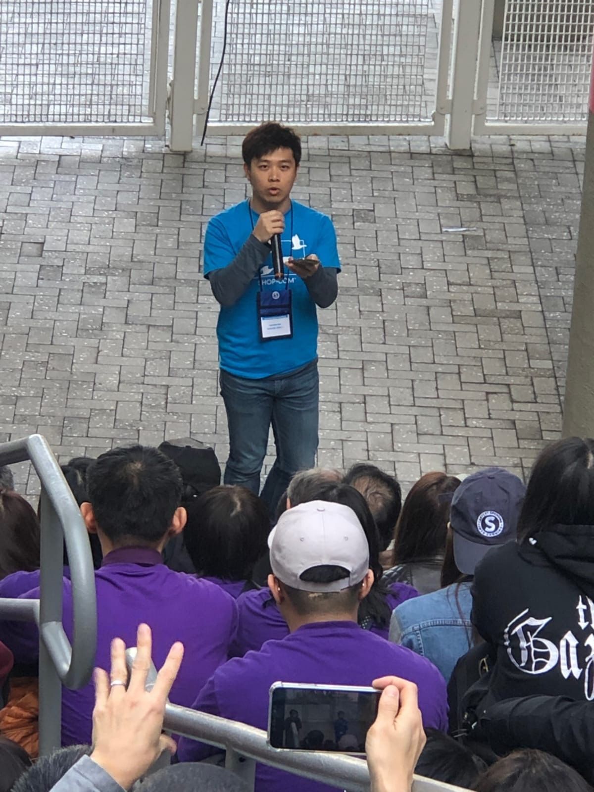 A man in a blue shirt speaks into a microphone outdoors. He gestures while addressing a crowd seated on steps.