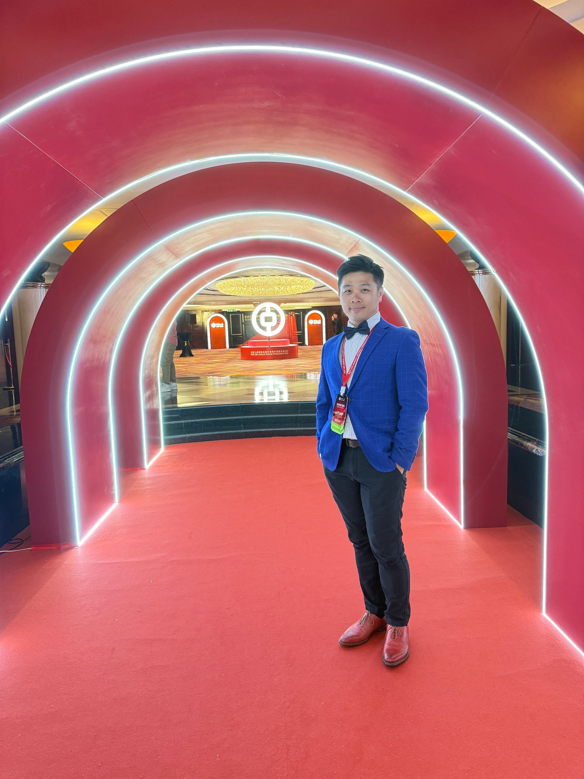 Man in blue blazer stands on a red carpet under a red and white archway; indoors.