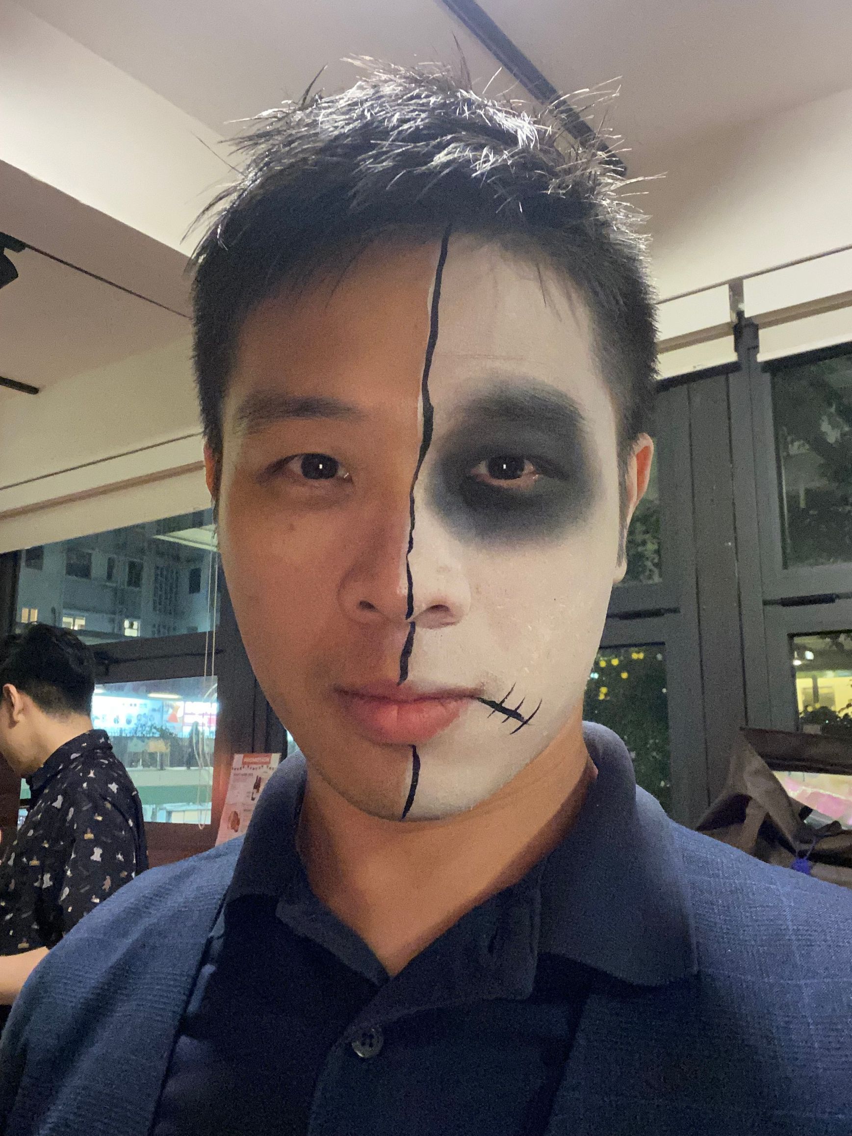 A person with half of their face painted as a skull for Halloween. One side is normal; the other is white with a black eye and stitched lines.