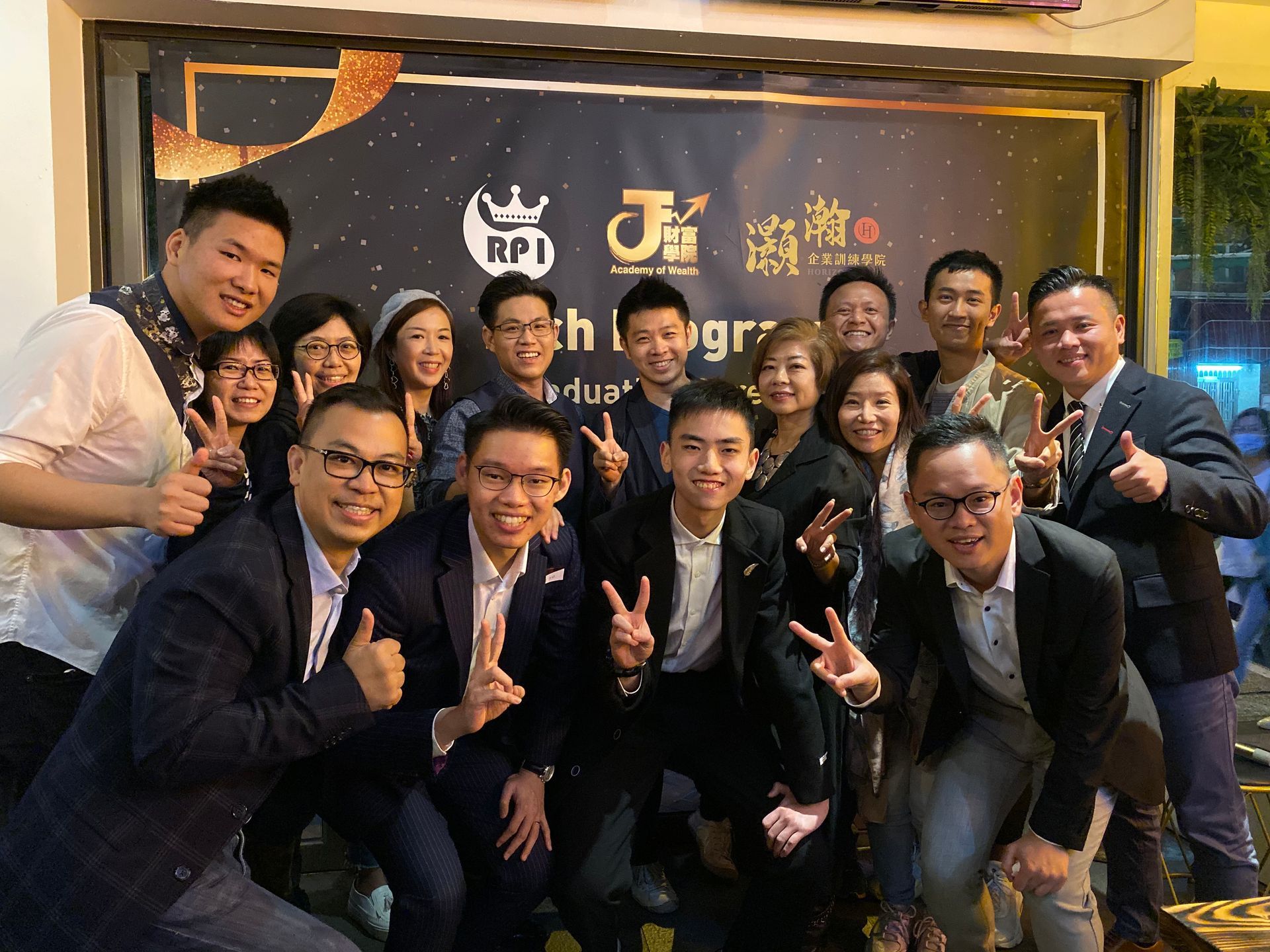 Group of people smiling, making peace signs, and celebrating. A banner with gold text and logos is in the background.