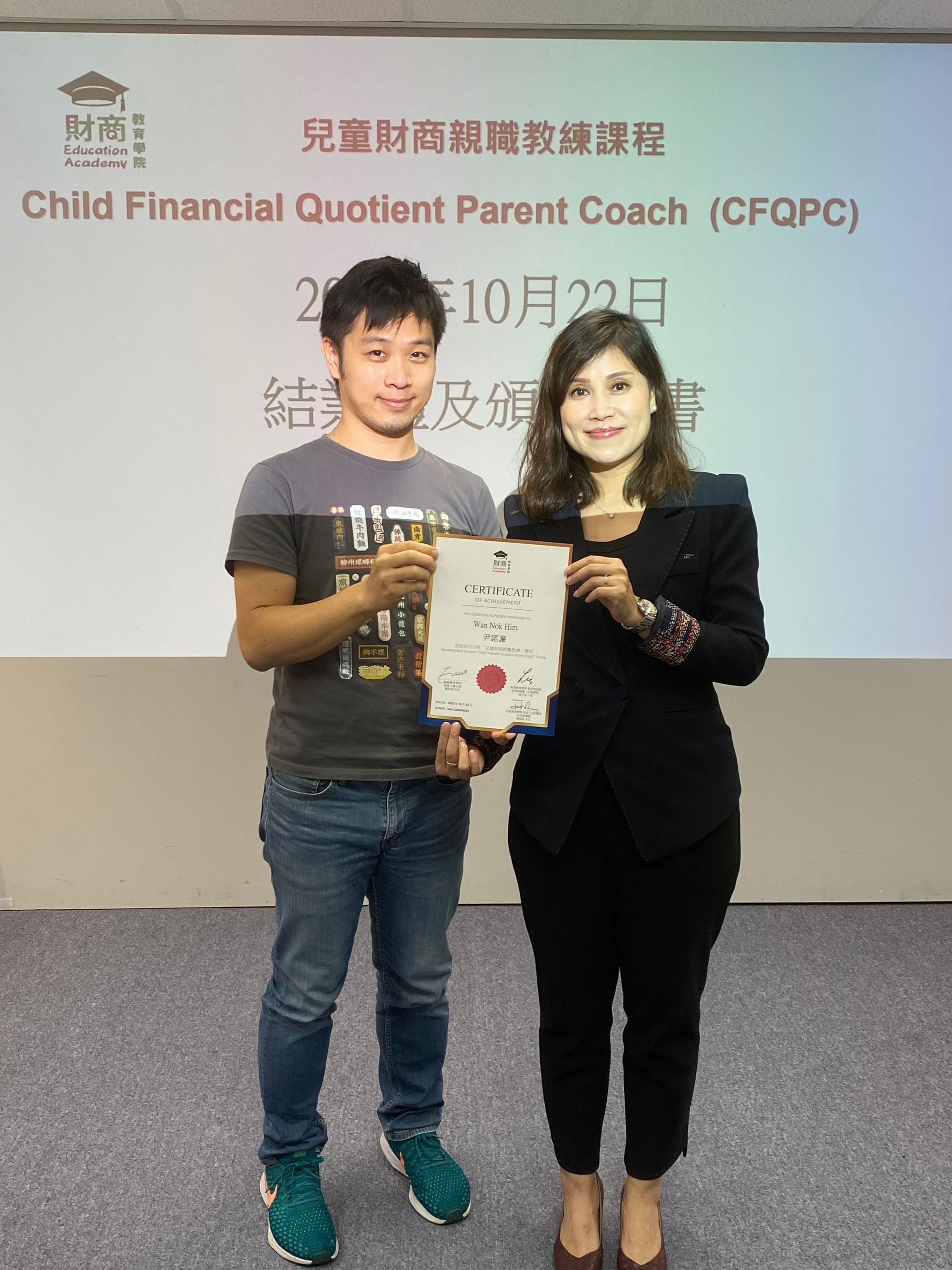 A man and woman holding a certificate, likely at a parent coaching event. The banner behind them reads