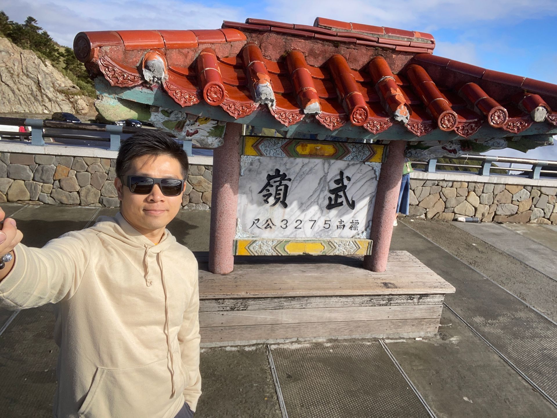 A man in sunglasses takes a selfie near a weathered, traditional sign with Chinese characters under a red-tiled roof.