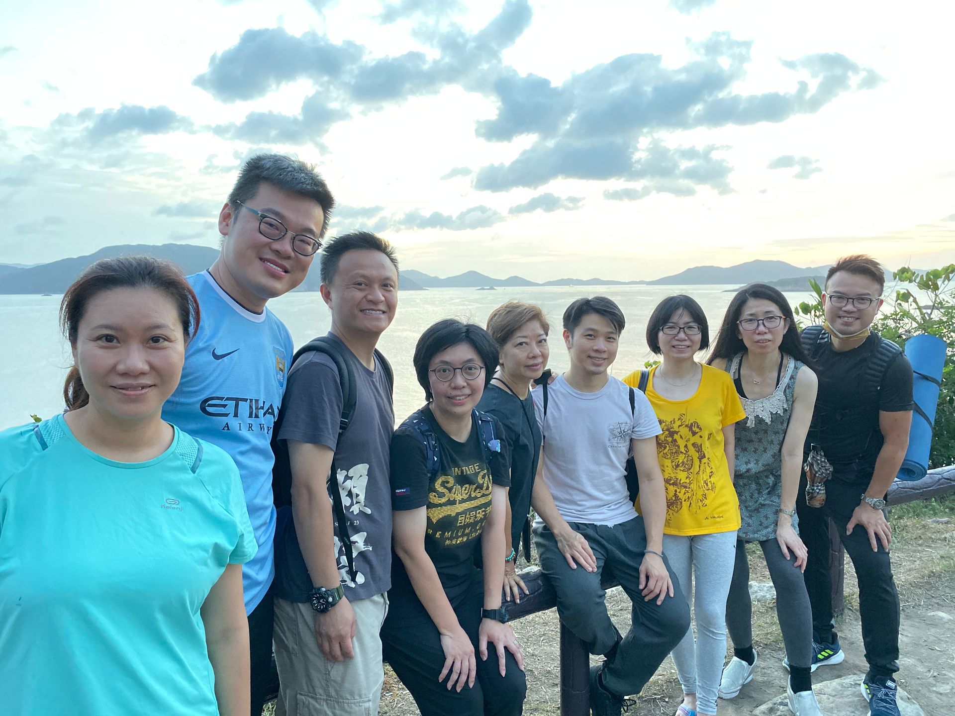 Group of ten people smiling at the camera in front of a body of water at dusk. The people are a mix of ages and ethnicities.
