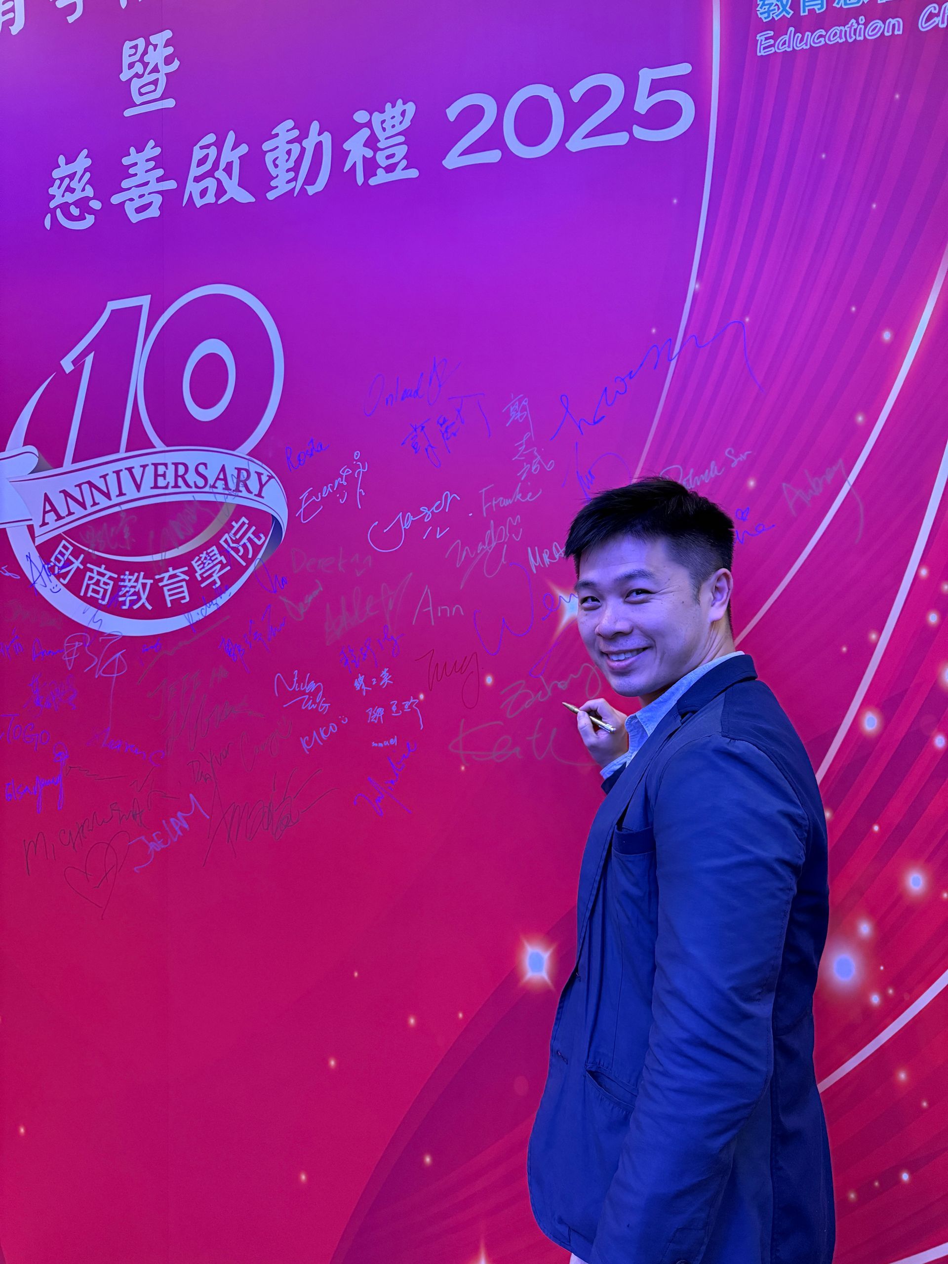 A man in a suit jacket smiles and points at a red wall with event details and signatures. A 10-year anniversary logo is visible.