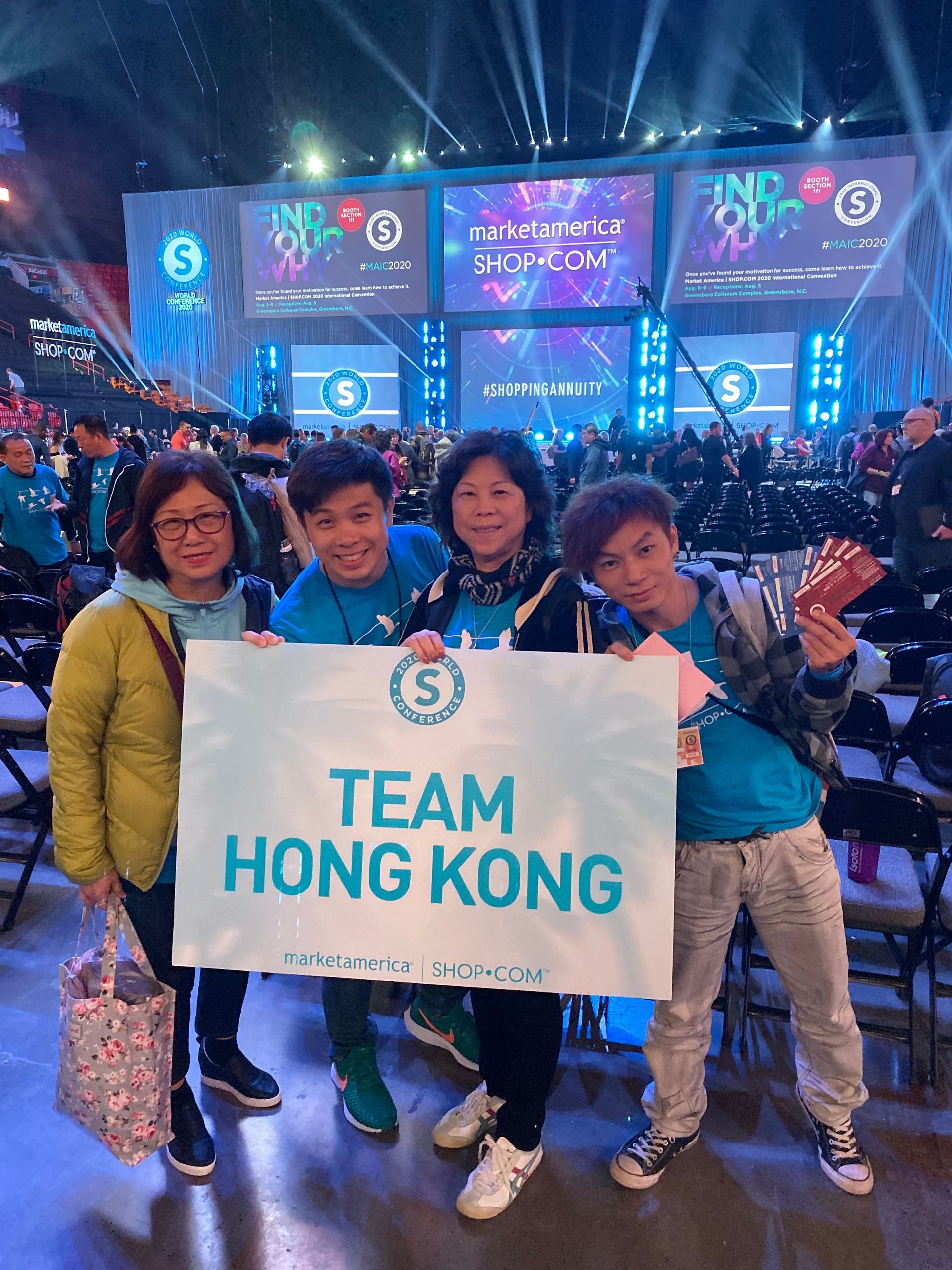 Four people from Team Hong Kong hold a sign at a large event with a stage and audience. They're smiling, wearing matching blue shirts.