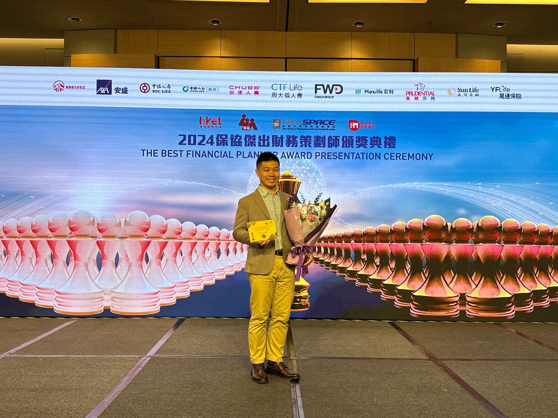 A man in a suit holds an award on a stage with a screen displaying event branding and lotus imagery.