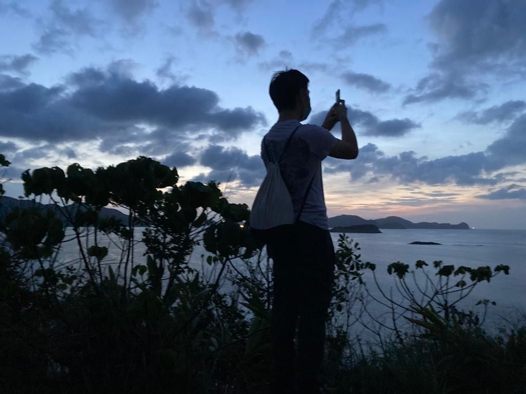 A person standing on a hill at dusk taking a photo of the ocean with a smartphone. The sky is cloudy with shades of blue and orange.