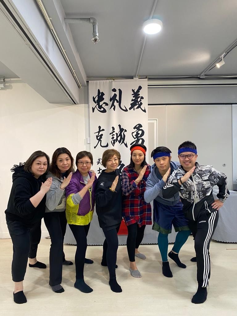 Group of eight people pose in front of a banner with Chinese characters. They wear casual clothing and make a karate chop gesture.