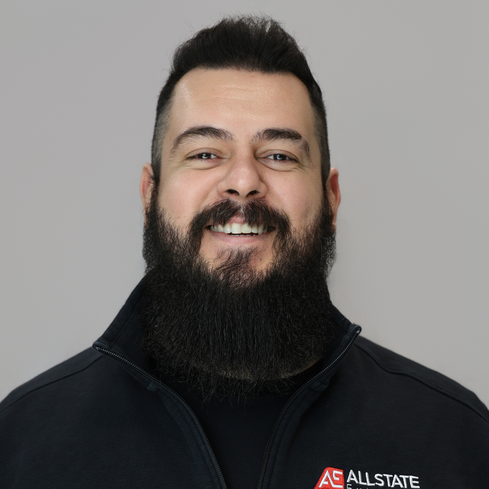 A man with a black hair and beard is wearing Allstate uniform in black color, smiling to the camera.