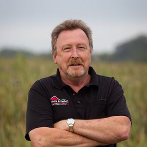 A man with a beard is standing in a field with his arms crossed.