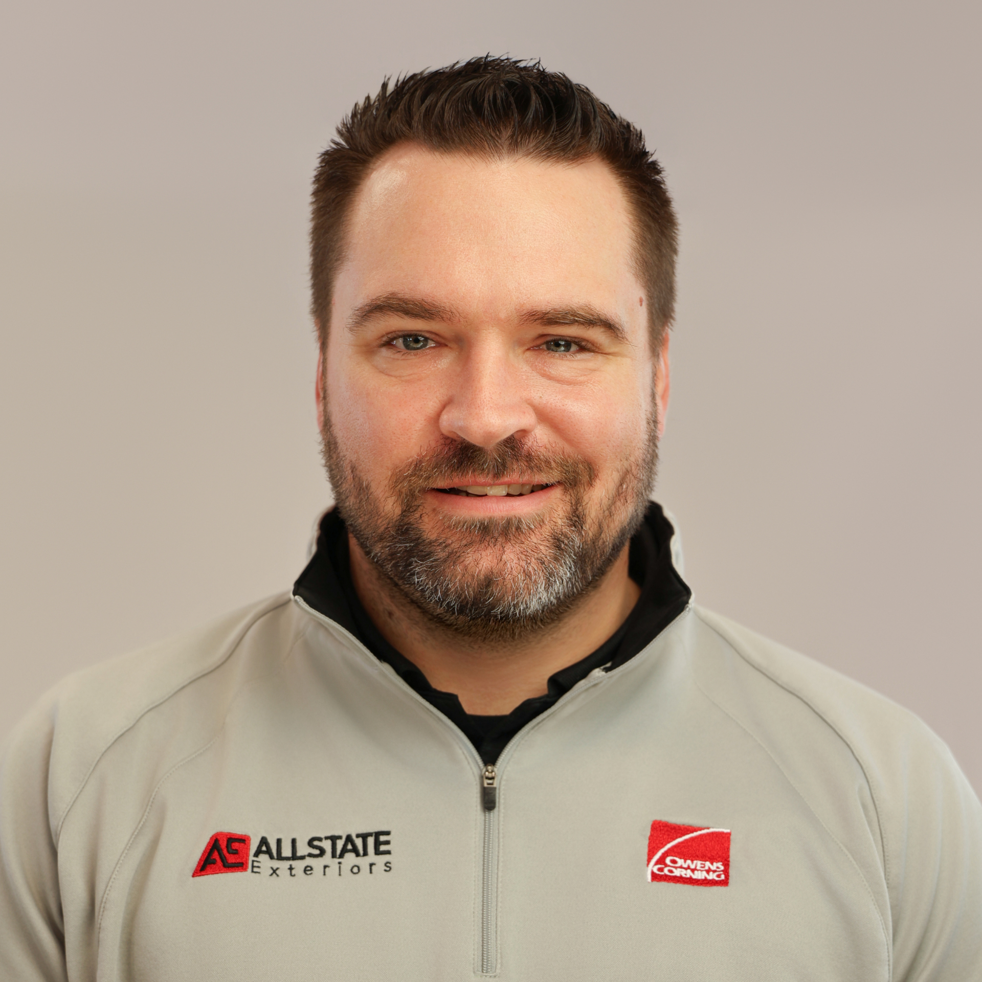 Man with arms crossed, smiling, wearing gray polo shirt, watch, and posing outdoors near tall grass.