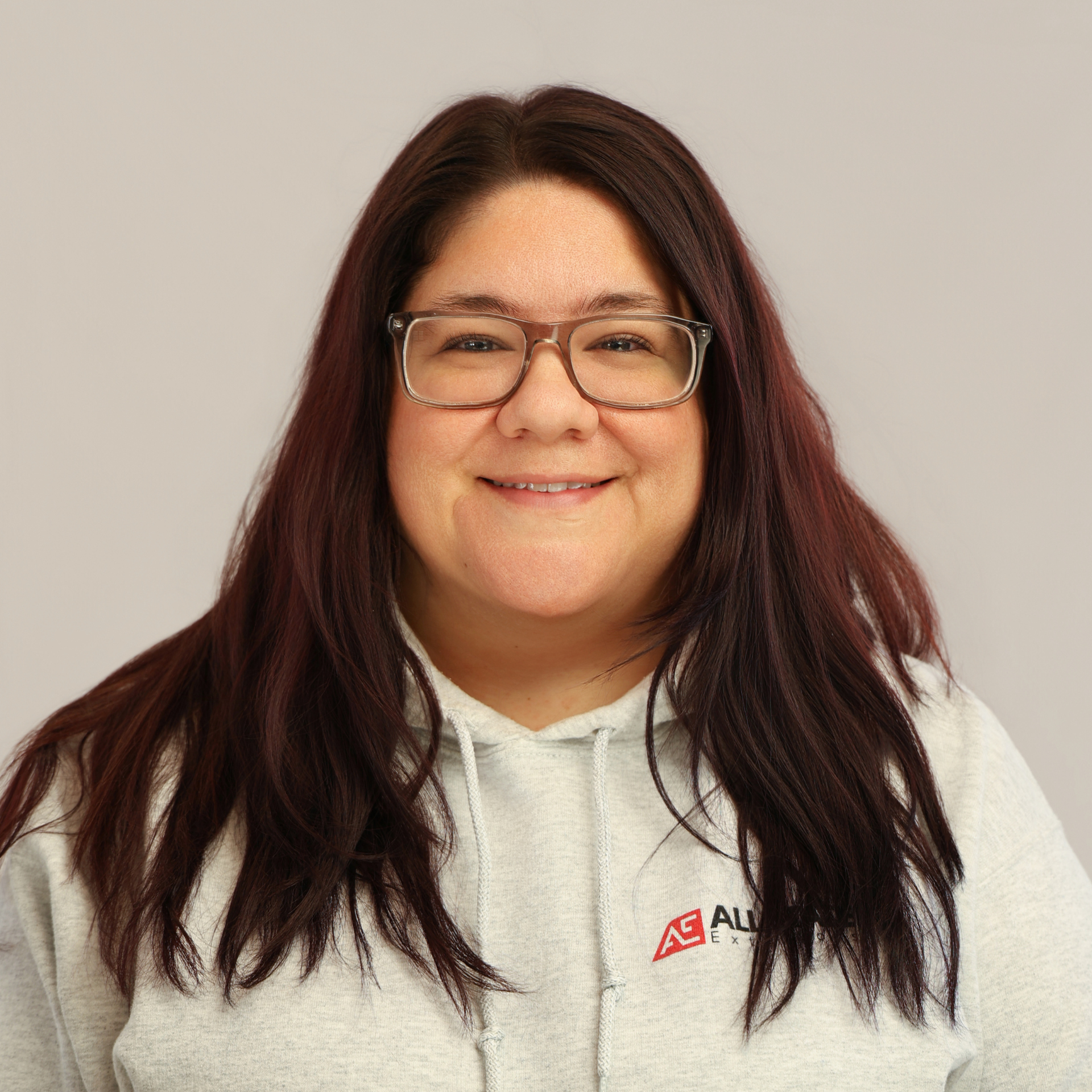 A woman wearing a gray sweatshirt and a necklace is smiling in front of a wooden wall.