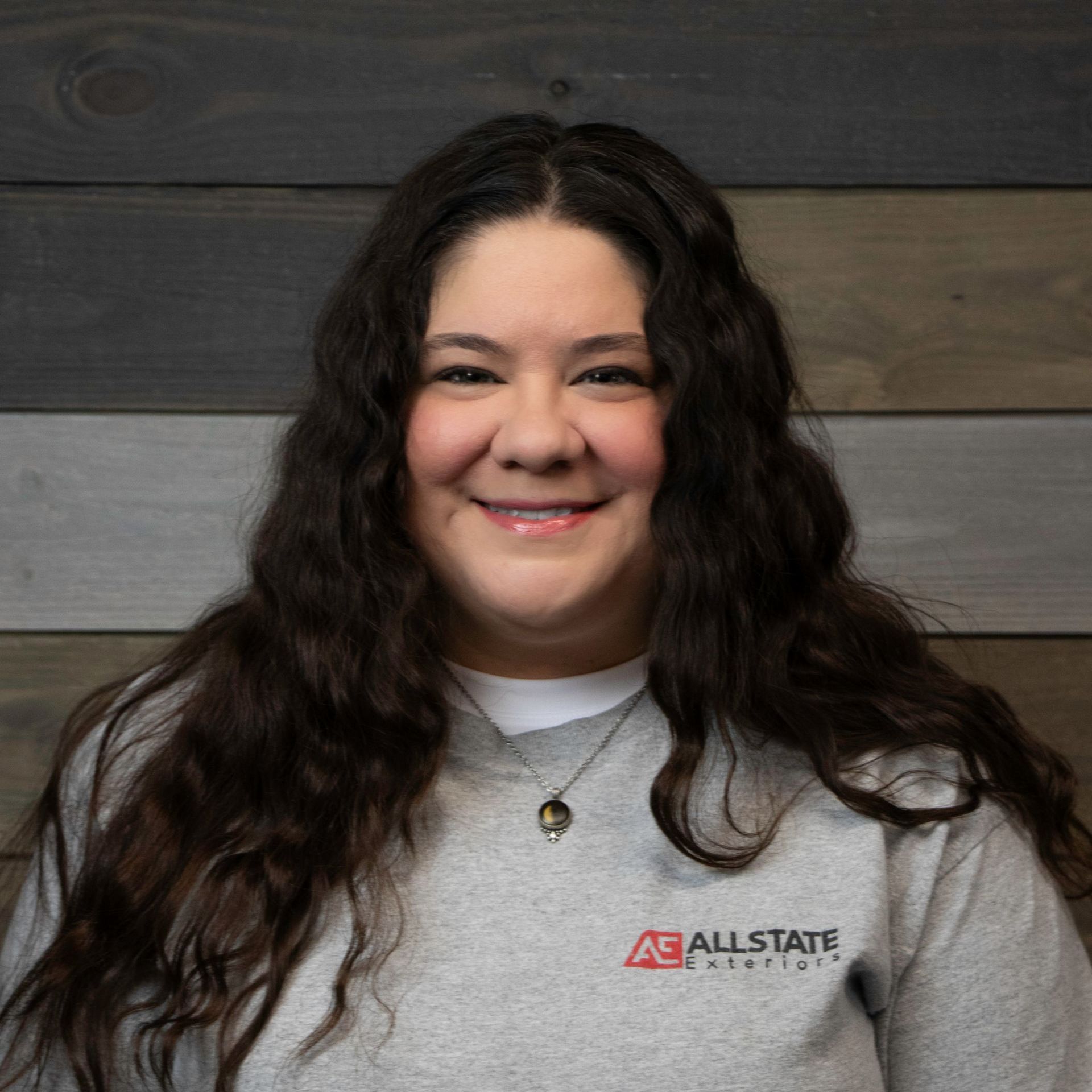 A woman wearing a gray sweatshirt and a necklace is smiling in front of a wooden wall.