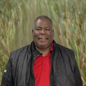 A man wearing a red shirt and a black vest is standing in front of a field of tall grass