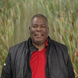 A man wearing a red shirt and a black vest is standing in front of a field of tall grass.