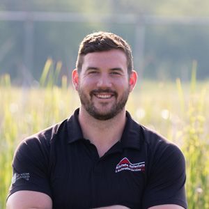 A man with a beard is standing in a field with his arms crossed and smiling.