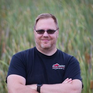 A man wearing sunglasses and a black shirt is standing in a field with his arms crossed.