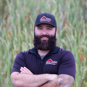 A man with a beard and a hat is standing in a field with his arms crossed.