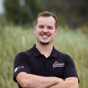 A man in a black shirt is standing in a field with his arms crossed and smiling.