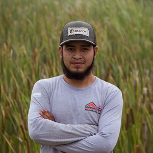 A man with a beard and a hat is standing in a field with his arms crossed.