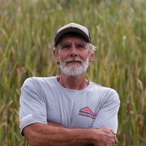 A man with a beard and hat is standing in a field with his arms crossed.