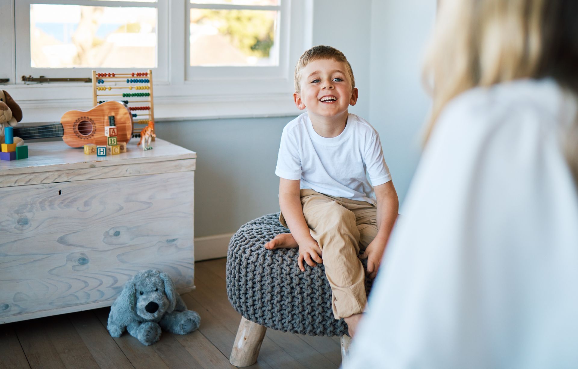 A young boy is sitting on an ottoman in a room talking to a woman.